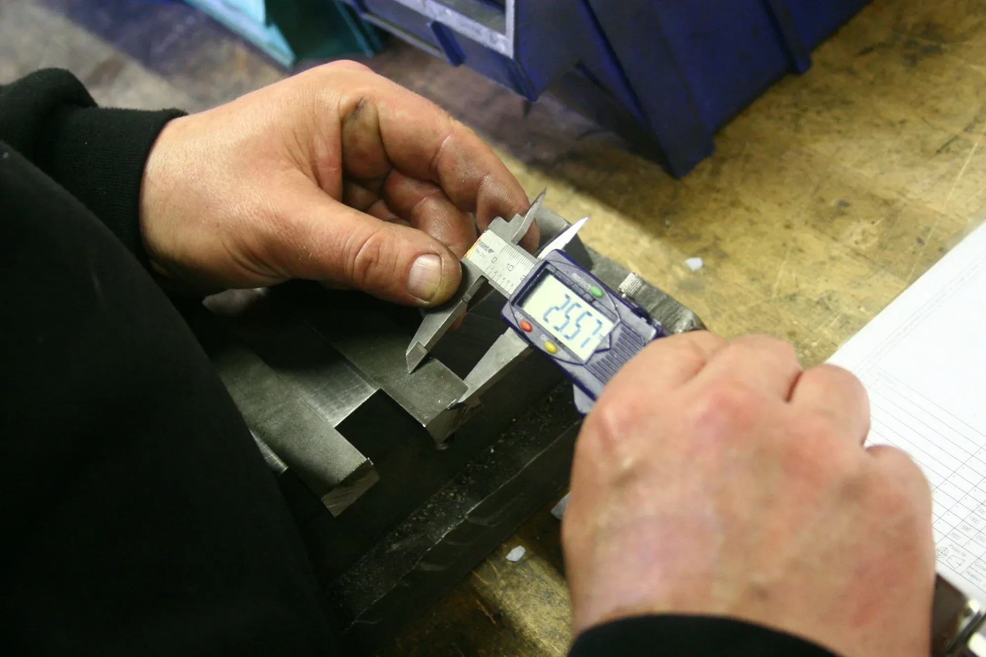 Hands measuring a machined part with digital callipers on a workshop bench.